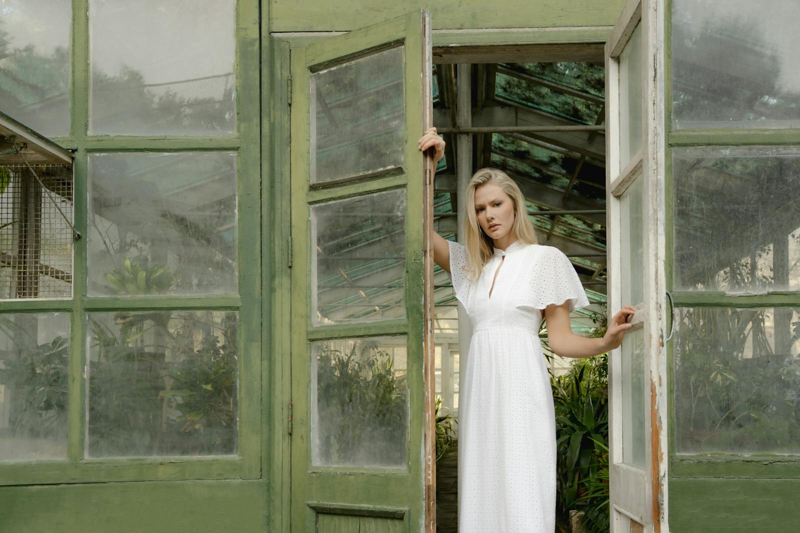 Stylish woman in a white dress stands at the entrance of a rustic greenhouse.